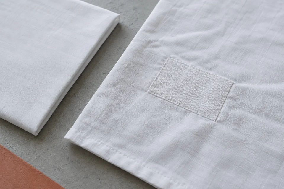 A neatly sewn white fabric patch on a sheet sample beside an unpatched reference on a grey surface