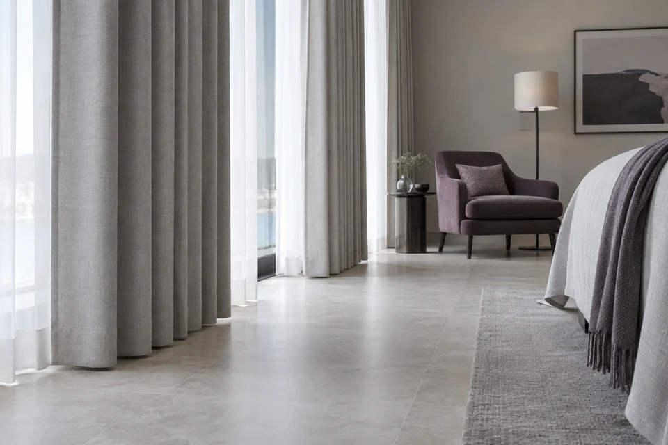 Grey curtains hang above a hotel room floor beside a plum armchair in soft daylight