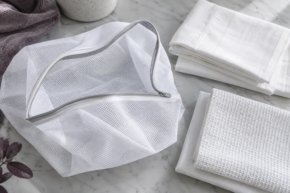 An open white mesh laundry bag containing folded linen rests on a pale grey worktable